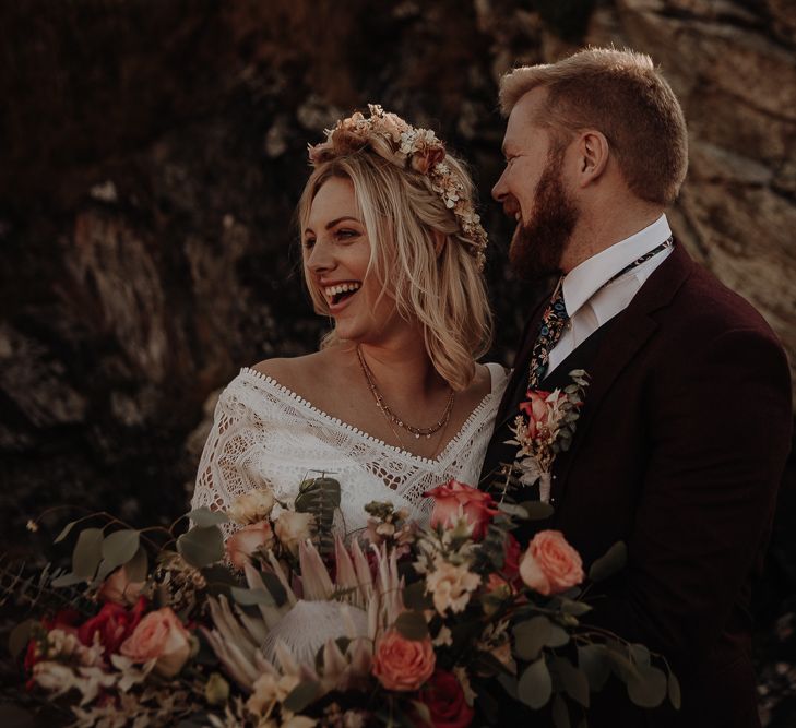 Large autumnal wedding bouquet held by bride in lace dress with flower crown and layered jewellery with groom 