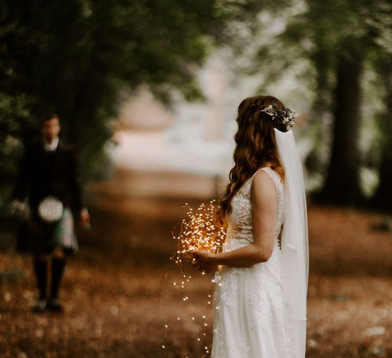 Bride in sparkly wedding dress holds string fairy lights in her hands as she awaits the groom