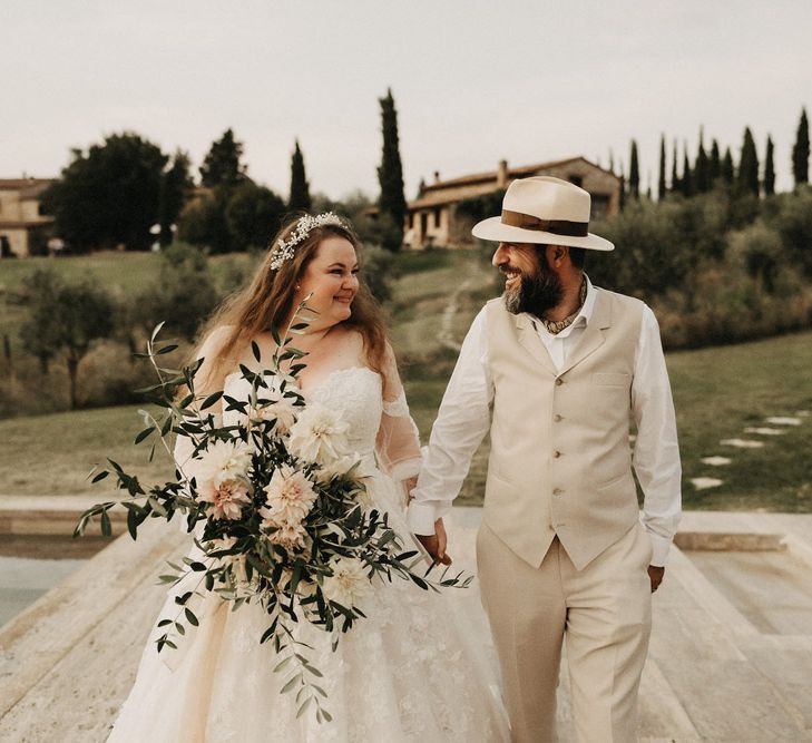 Bride and groom smiling, holding hands with an olive bouquet