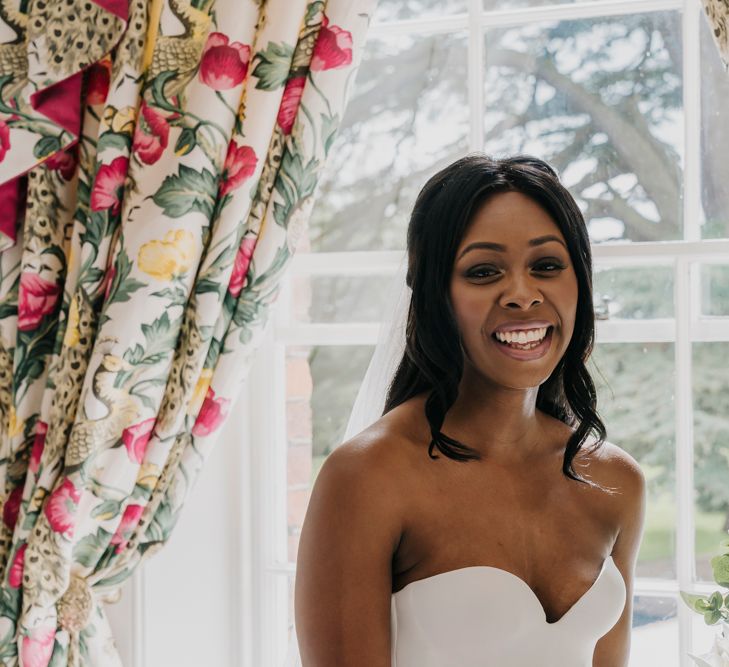 A Black bride with long loose curls stands in front of a window and smiles to camera