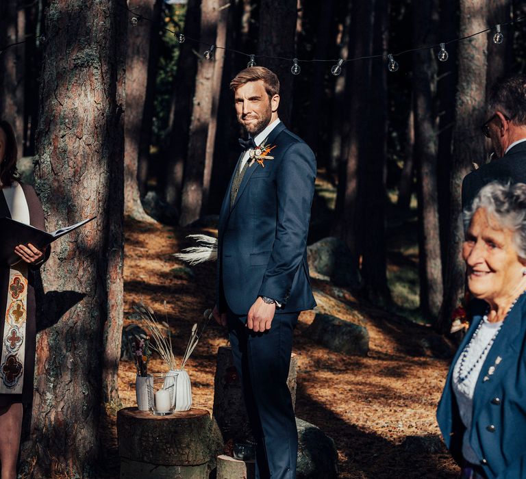 Grooms awaits bride during outdoor ceremony with strung lights in the background