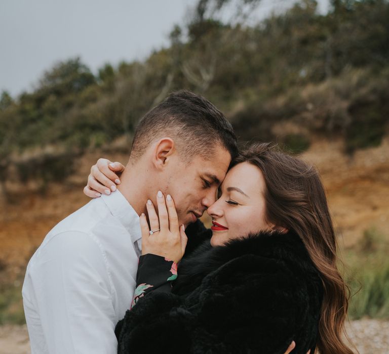 Intimate engagement session photograph with bride-to-be with beige nail polish caressing her groom-to-be's face showing off her diamond engagement ring 