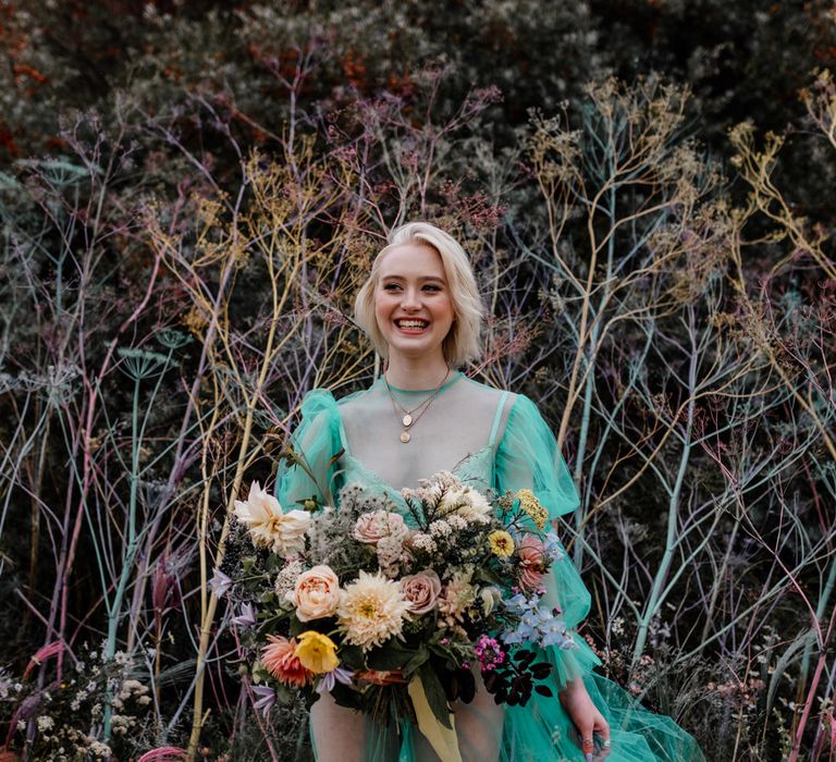 White bride with short blonde hair in a sheer pastel wedding dress on the beach holding an oversized colourful bouquet tied with ribbons