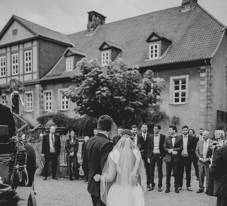 Bride in white Jesus Piero gown and cathedral length veil arrives at German wedding reception with groom in black Tiger of Sweden tux