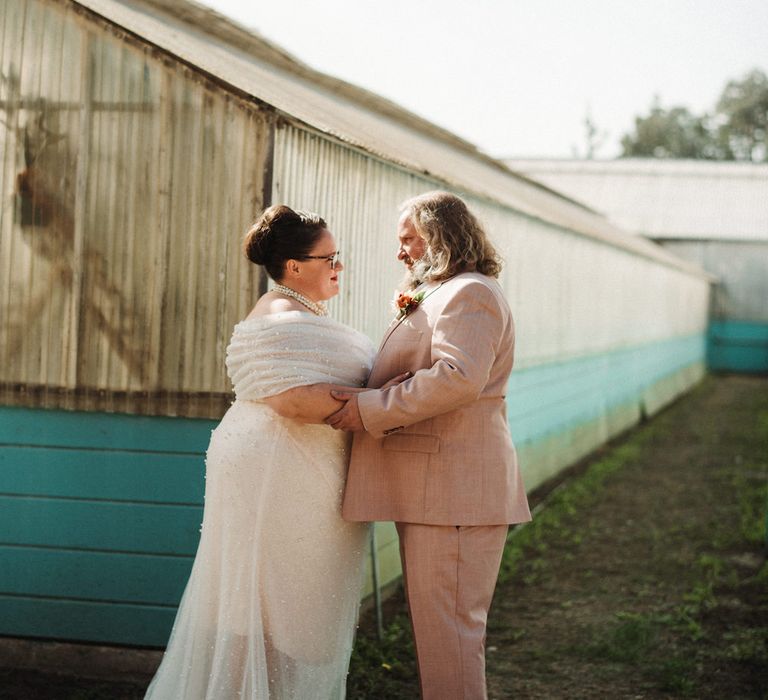 Bride with a top knot in an off the shoulder wedding dress embracing her husband in a pink suit 