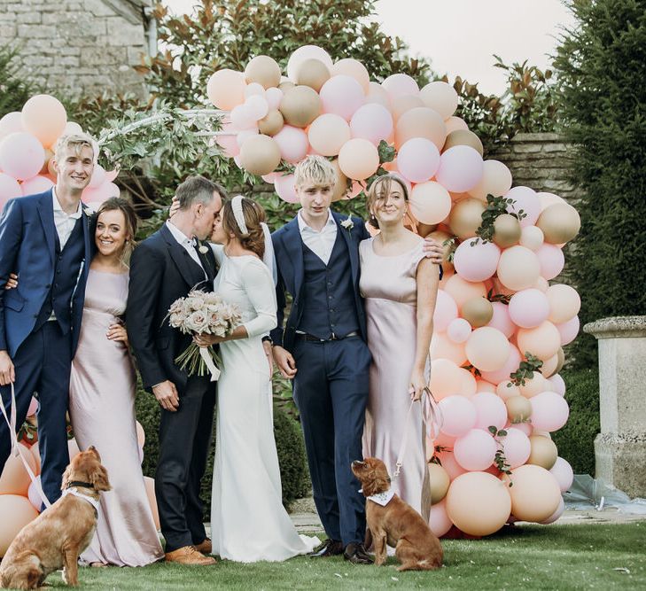 Bride in Elbeth Gillis gown kisses groom in navy Cad & the Dandy suit in front of pastel pink and gold balloon arch with groomsmen and bridesmaids at Euridge Manor wedding