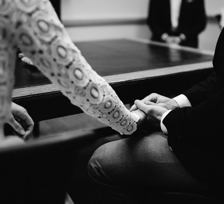 Bride sat in lace long sleeved Self Portrait wedding dress holds hand of groom in brown woollen blazer and grey trousers at Bristol Registry Office wedding