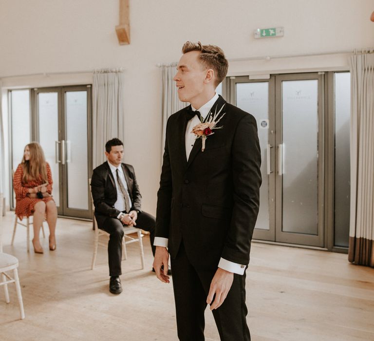 Groom's reaction to his bride's entrance in a black tuxedo and red flower buttonhole with pampas grass 