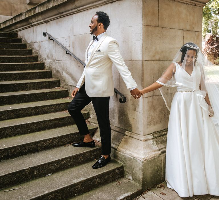 Stylish groom in a white tuxedo jacket and bow tie standing behind a wall holding hands with his bride in a princess wedding dress and cathedral length veil 