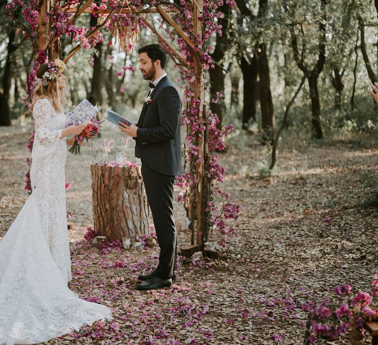 Bride in long sleeved lace boho wedding dress and flower crown reading does with groom in navy suit and bow tie in front of wooden wedding arch decorated with pink flowers