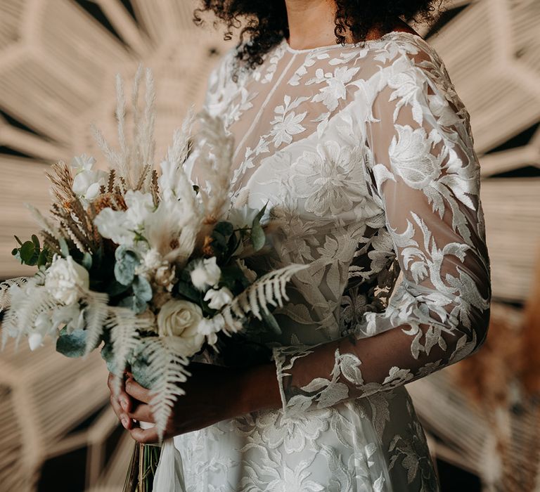 Bride looks at the camera whilst holding white floral bouquet and standing in front of Macrame decor