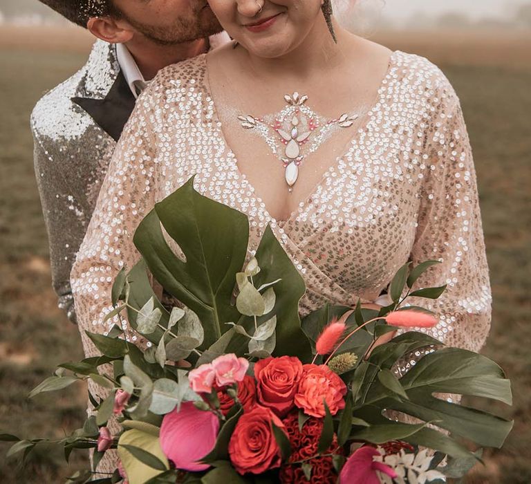 Bride in gold V neck long sleeved sequin wedding dress and face gems holds red rose and foliage bouquet whilst groom in silver sequin suit jacket hugs her from behind