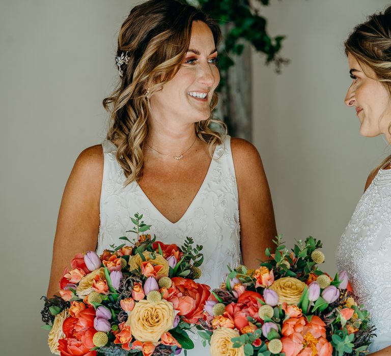 Brides look lovingly at one another on their wedding day whilst holding bright floral bouquets