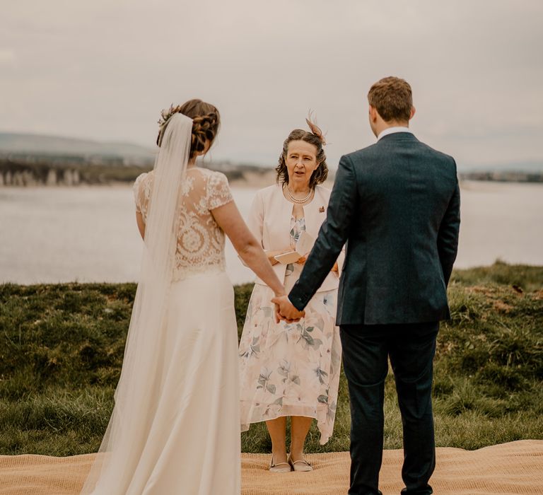 Bride in lace top with capped sleeves, veil and satin skirt holds hands with groom in navy suit for clifftop ceremony at Dunluce Castle wedding