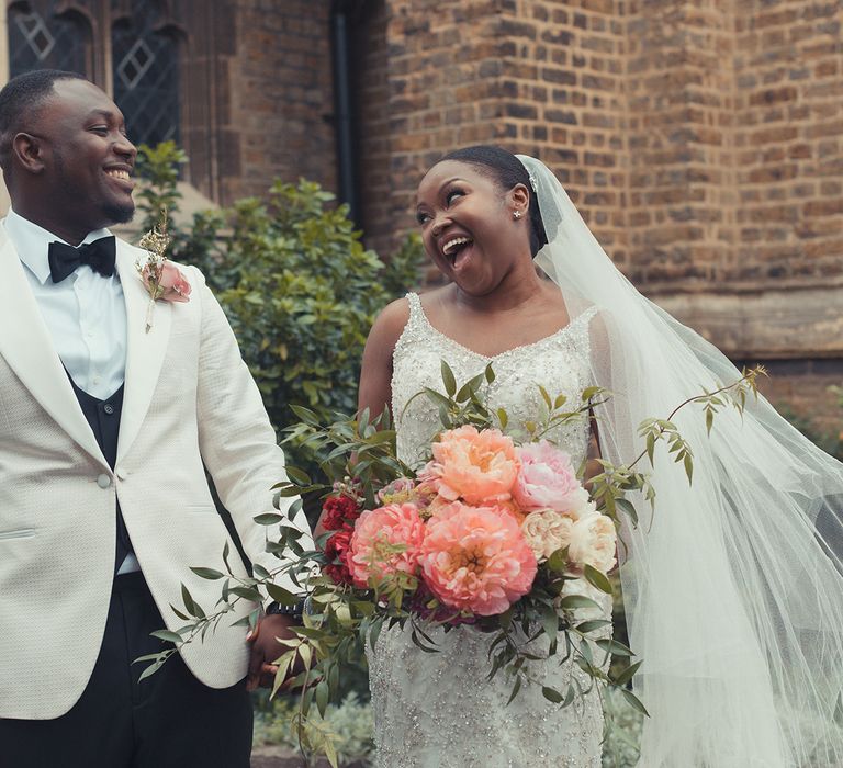 Bride & groom stand outdoors as groom wears white tuxedo and bride holds brightly coloured peach bouquet