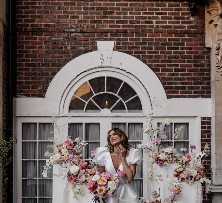 Beautiful bride in a stylish wedding dress with front split holding a pink peeled back rose bouquet standing in front of blush floral arrangements 