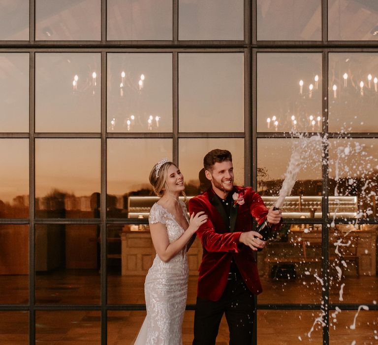 Bride in a fitted shimmering wedding dress and groom in a red velvet tuxedo popping champagne at Botley Hill Barn 