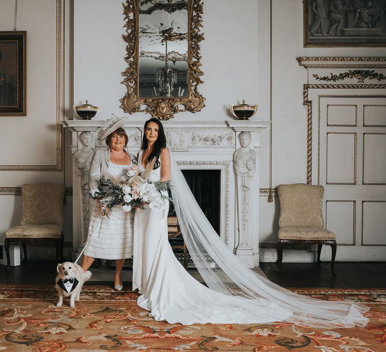 Bride in white Pronovias wedding dress with train and veil holds large white, pink and green wedding bouquet as she stands with wedding guest in cream dress and hat holding white dog on a lead in a formal room at Came House Dorset