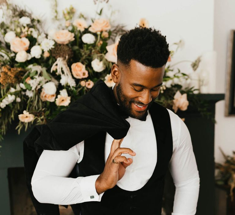 Groom smiling and looking down as he holds his black velvet suit jacket over his shoulder