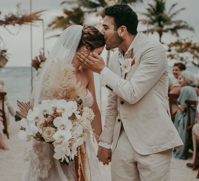 Groom kisses bride on the forehead as they hold hands and walk down the aisle after their beach ceremony wedding