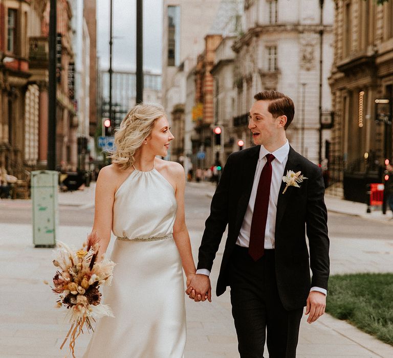 Bride in a Kate Beaumont champagne coloured wedding dress holding hands with her groom in a black suit and burgundy tie at their Leeds city wedding 