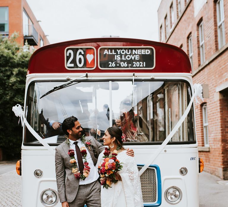 A bride and groom stand in front of a white open top bus.