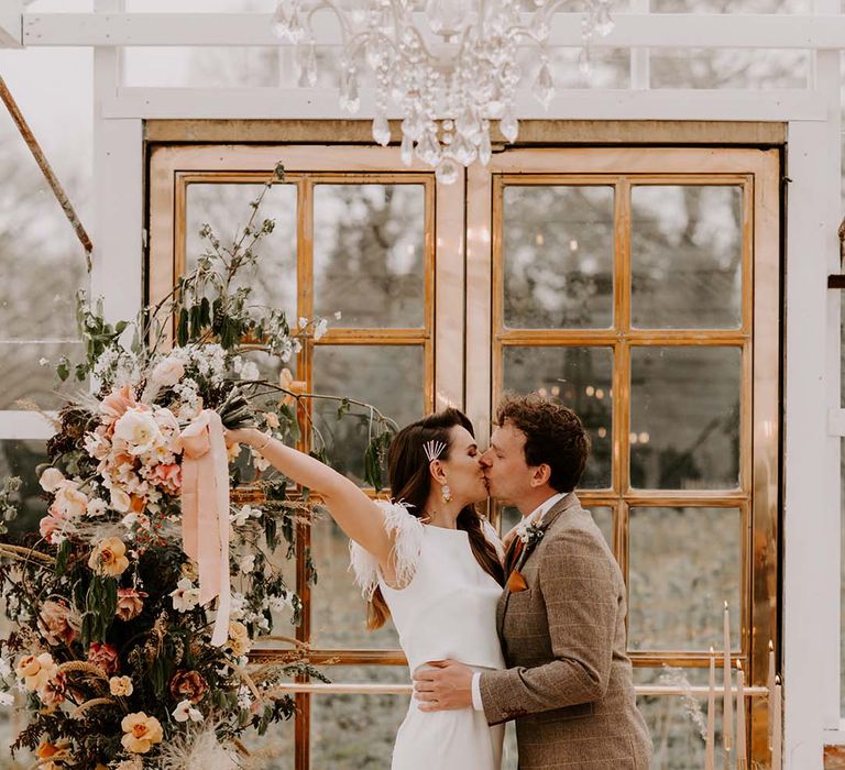 Groom in a brown check suit kissing his bride in a fitted wedding dress with feather cap sleeves at the altar decorated in autumnal fresh and dried flowers 