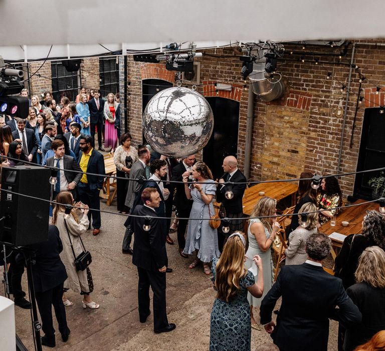 Wedding guests stand in exposed brick courtyard with wooden tables and disco ball before wedding at Loft Studios London