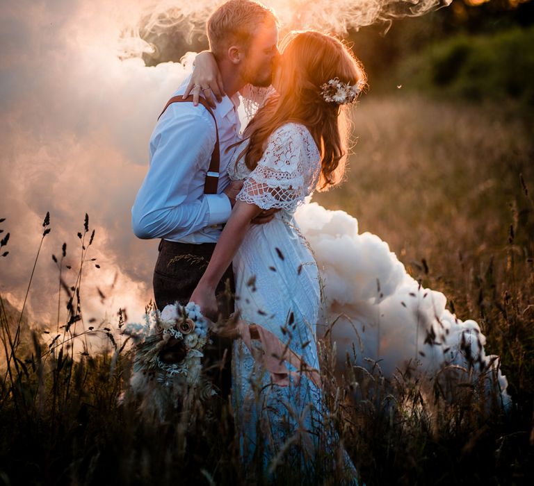 Boho wedding with bride in a lace wedding dress holding a dried flower bouquet kissing her grooming a navy military uniform during golden hour as a white smoke grenades blows in the wind