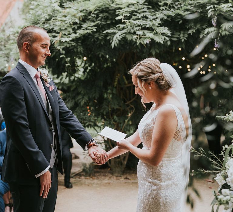 Groom in dark three piece suit with grey waistcoat and pink tie holds hands with bride in lace Justin Alexander wedding dress and veil as she reads vows during outdoor wedding ceremony at Tythe Barn