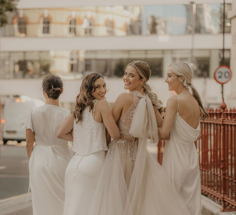 Fun bridal party portrait with them looking back over their shoulders. The bride wears a tulle wedding dress with bow, and the bridesmaids satin dresses