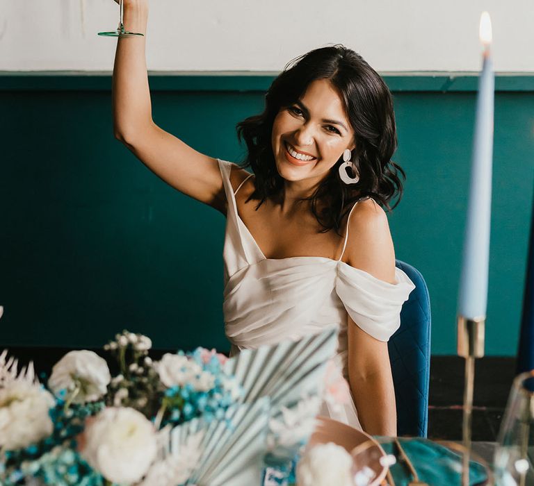 Bride with short wavy hair in a cold shoulder wedding dress holding her coloured glass in the air at her teal wedding 