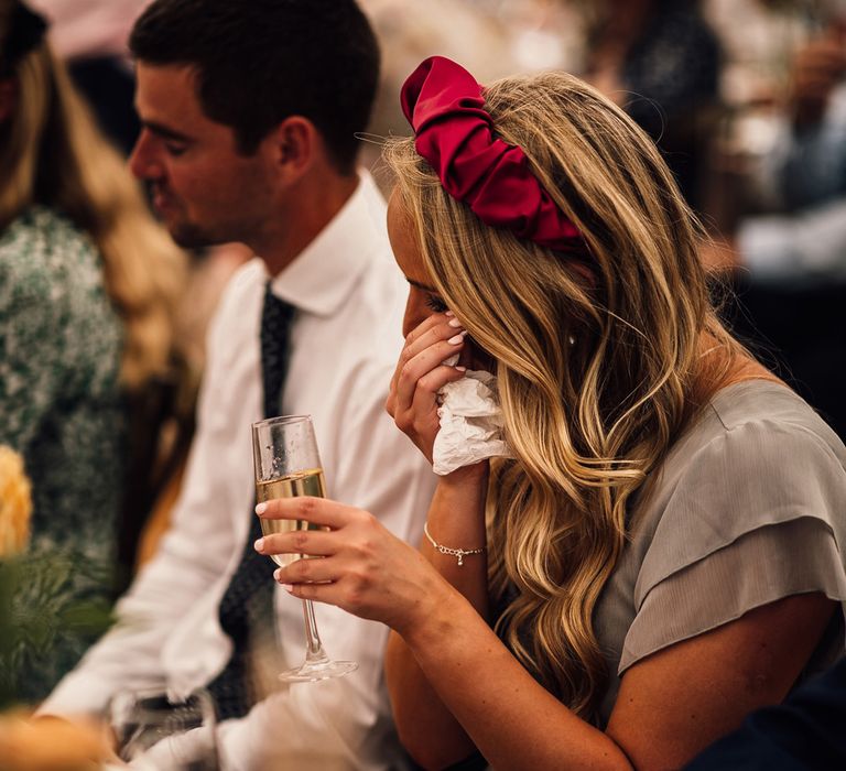 Bridesmaid in grey ruffled bridesmaid dress and pink headband wipes eyes with tissue as she holds glass of Prosecco at garden wedding reception in Cornwall