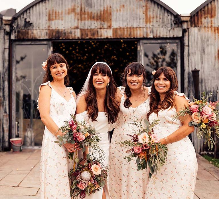 Bridesmaids in cream floral sating bridesmaid dresses posing with the bride holding pink protea wedding bouquets 