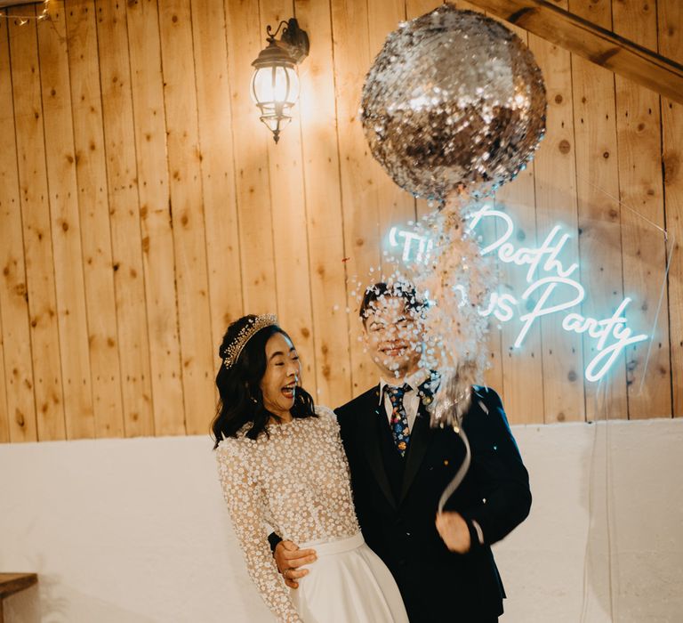 Groom in dark blue suit with floral tie stands with bride in long sleeved applique wedding dress and headpiece as they pull disco ball piñata at rustic wedding ceremony