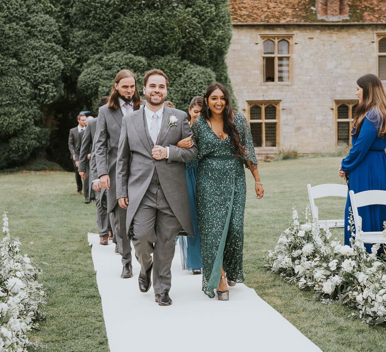 Groomsmen in morning suits walk up the white carpeted aisle with bridesmaids in green bridesmaid dresses during outdoor wedding ceremony at Notley Abbey