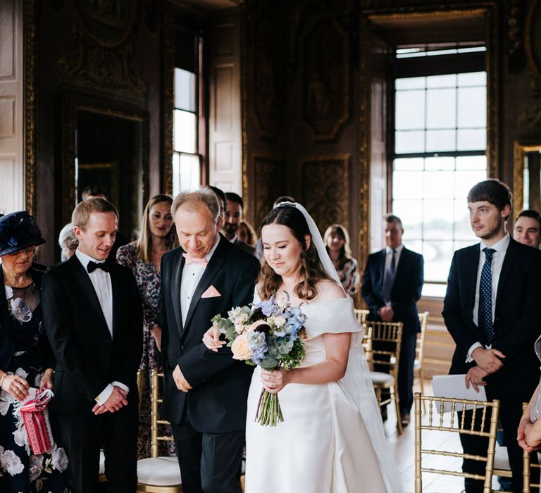 Bride walks down the aisle whilst holding pastel floral bouquet with her father