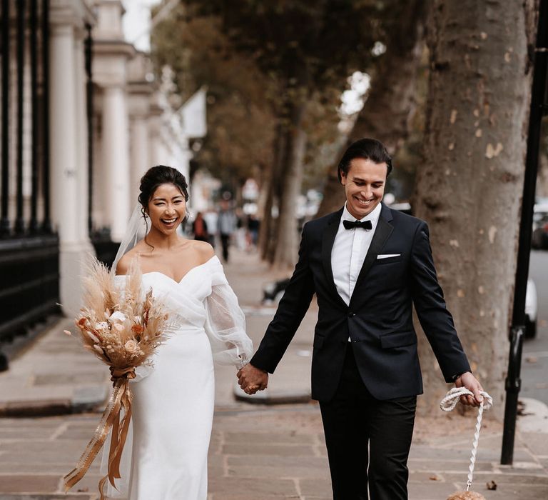 Bride in a long sleeve wedding dress holding hands with her groom in a tuxedo as they was their dog to their surprise Town Hall wedding ceremony 