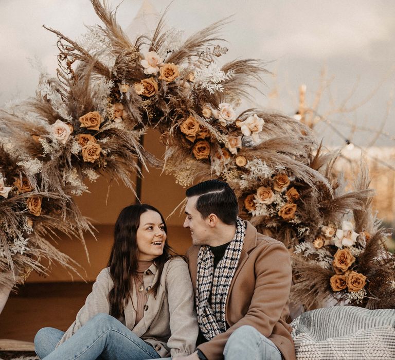 Groom-to-be in jeans and a camel coat embracing his fiancé in front of a bell tent decorated with dried and fresh flower decor