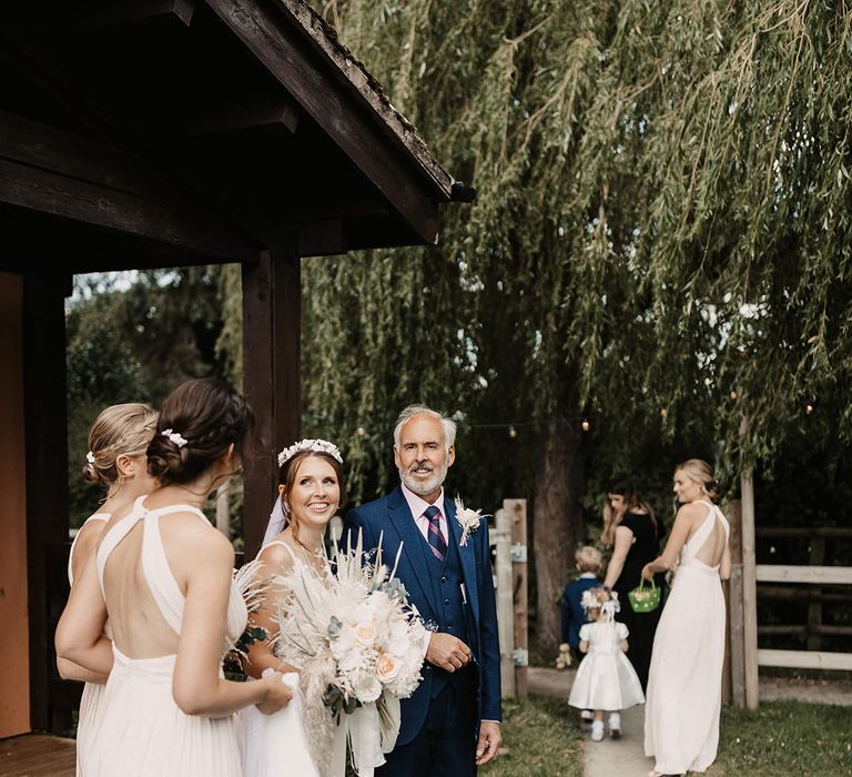 Bride holds her fathers arm and looks toward her bridesmaid outdoors on her wedding day