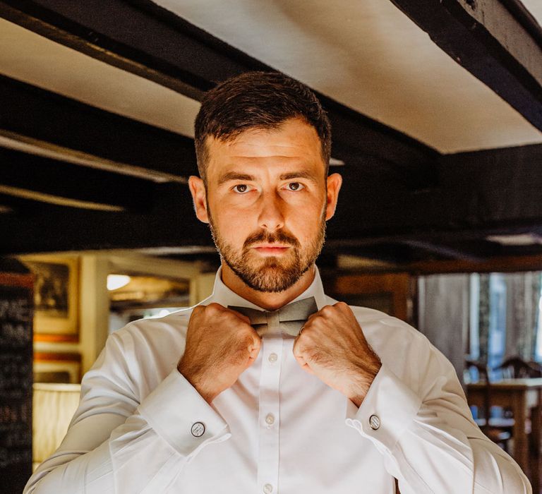 Groom in brown tweed trousers and belt, white shirt and tweed bow tie before wedding ceremony