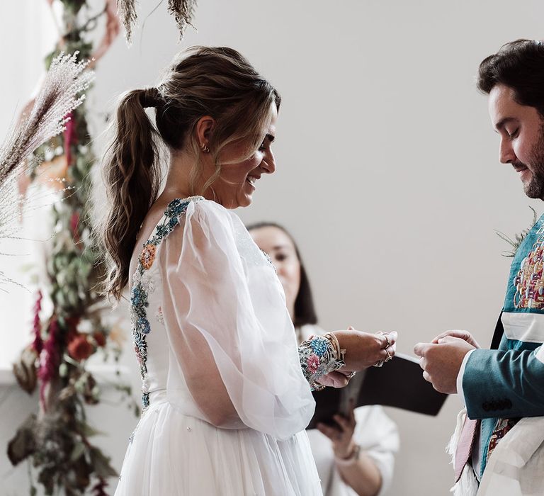 Bride with ponytail and mesh sleeve wedding dress exchanges rings with groom in red kippah and tallit during wedding ceremony at Iscoyd Park