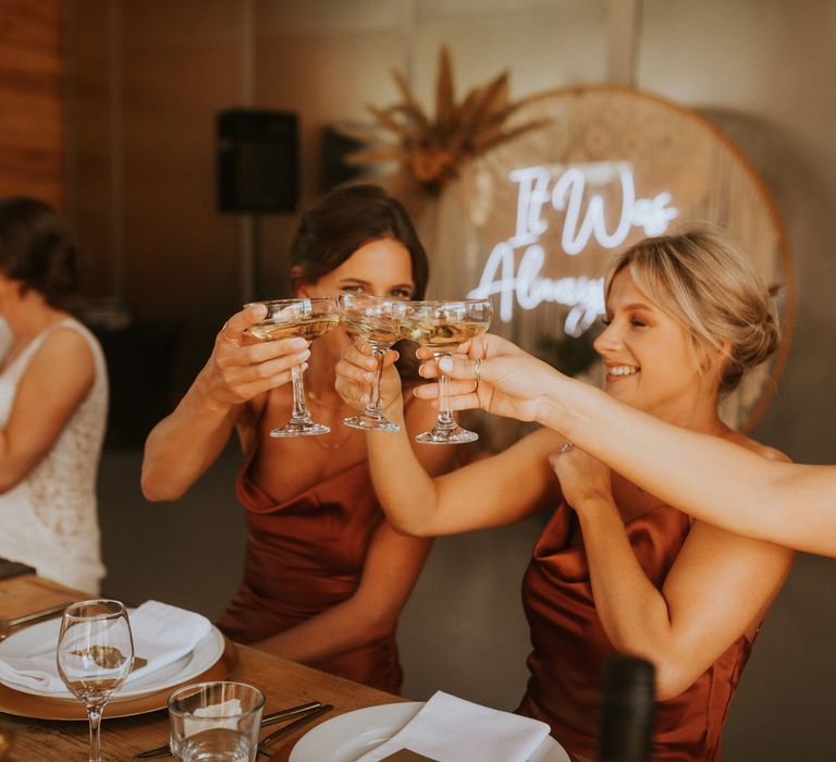 Bridesmaids in orange satin cowl neck bridesmaid dresses raise their champagne coupes during barn wedding reception