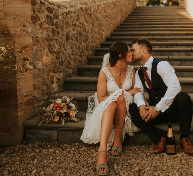 Bride in lace wedding dress and green block heels with bow detail sits on stone steps kissing from in white shirt and waistcoat 