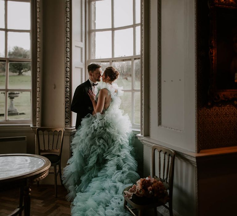 Bride in a light blue ruffle wedding dress by Millia London standing in the window at Newburgh Priory with her groom in a tuxedo 