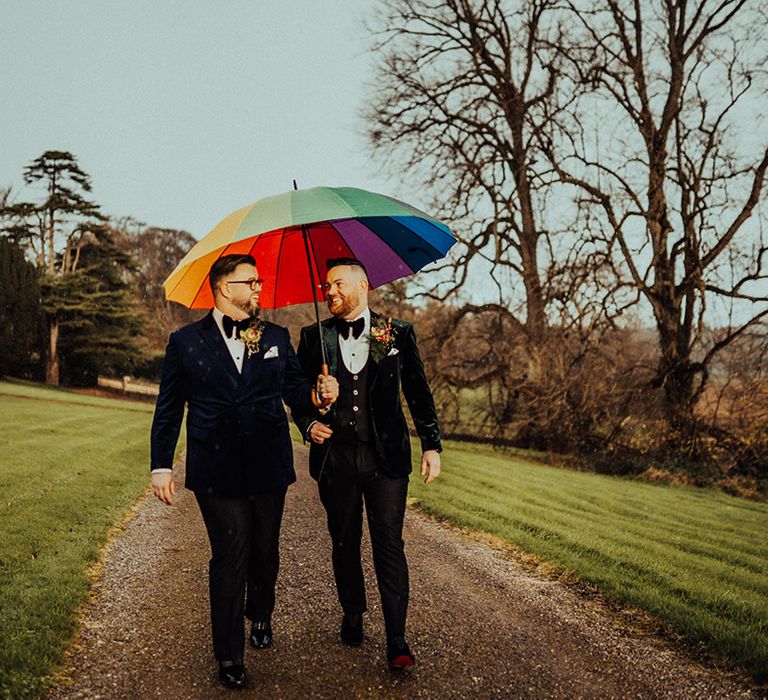 Grooms walk under rainbow coloured umbrella in velvet suit jackets for their wedding