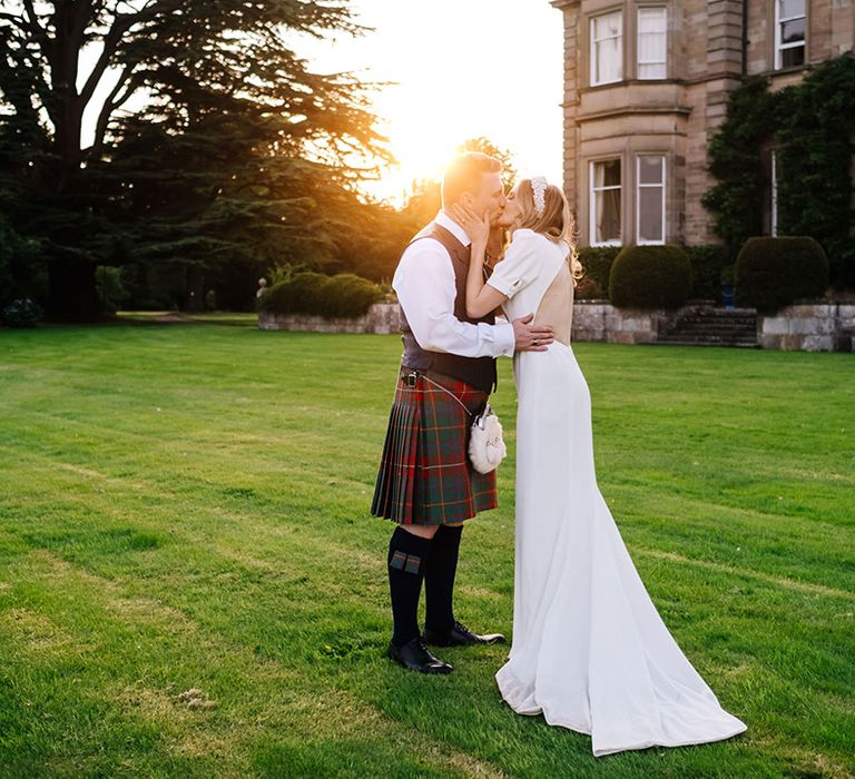 Bride in cutout back wedding dress with puff sleeves and pearl headband kisses groom in traditional kilt and blue waistcoat 