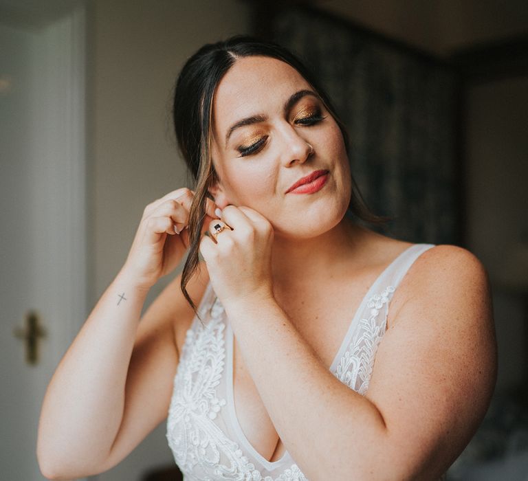 Bride in v-neck lace wedding dress and gold eyeshadow puts in her earrings for her wedding day