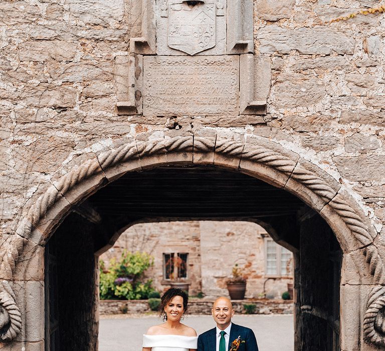 Groom in blue suit and green tie with bride in off the shoulder wedding dress stand under archway