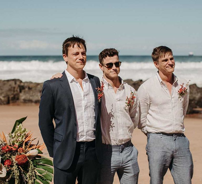 Groomsmen in shirts and light blue trousers and red tropical buttonholes match the groom's in suit jacket for beach wedding